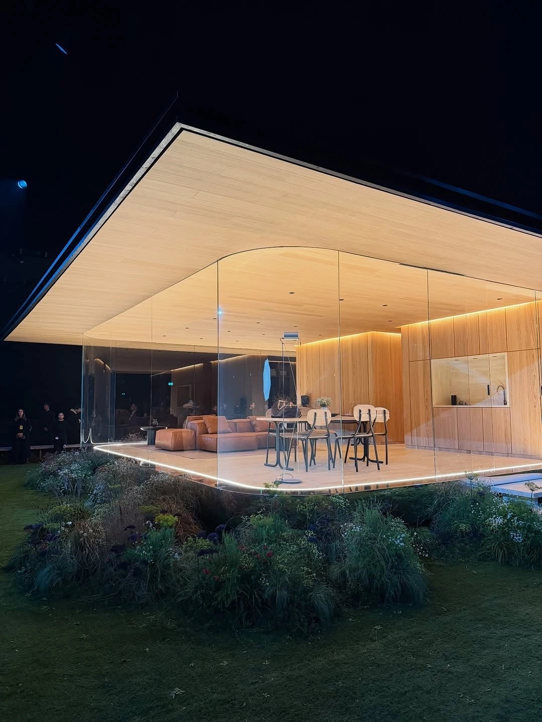 Glass pavilion at night, timber ceiling glowing over the garden.